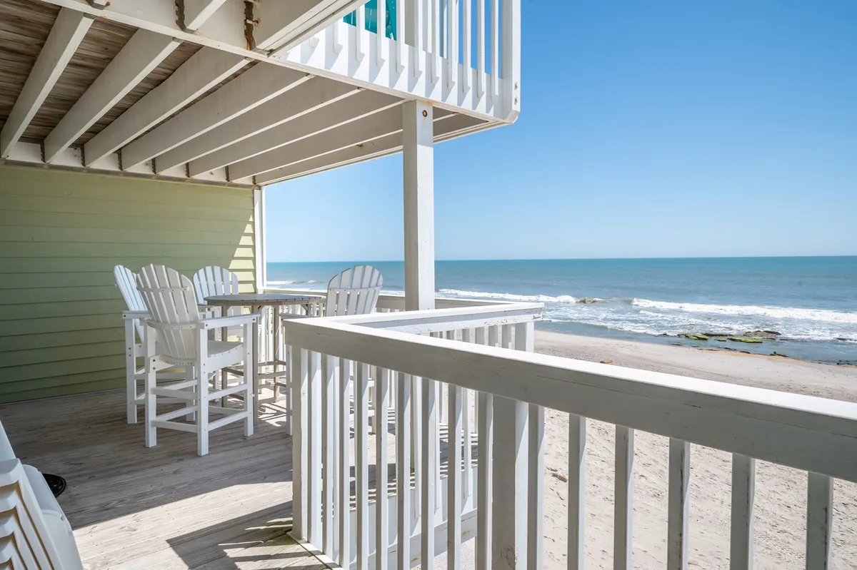 A serene beachfront view with outdoor seating overlooking gentle ocean waves. The foreground features a comfortable arrangement of chairs and a table, perfect for relaxing and socializing while enjoying panoramic ocean vistas. The calm sea and soft sky create a tranquil, inviting atmosphere.