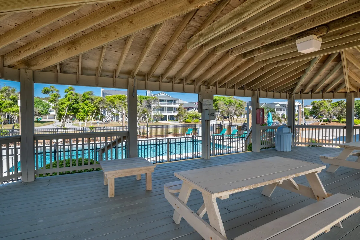 A vibrant outdoor scene featuring wooden picnic tables set beside a sparkling swimming pool, surrounded by lush greenery. The inviting space is ideal for family gatherings, picnics, and afternoons spent socializing and relaxing by the pool.
