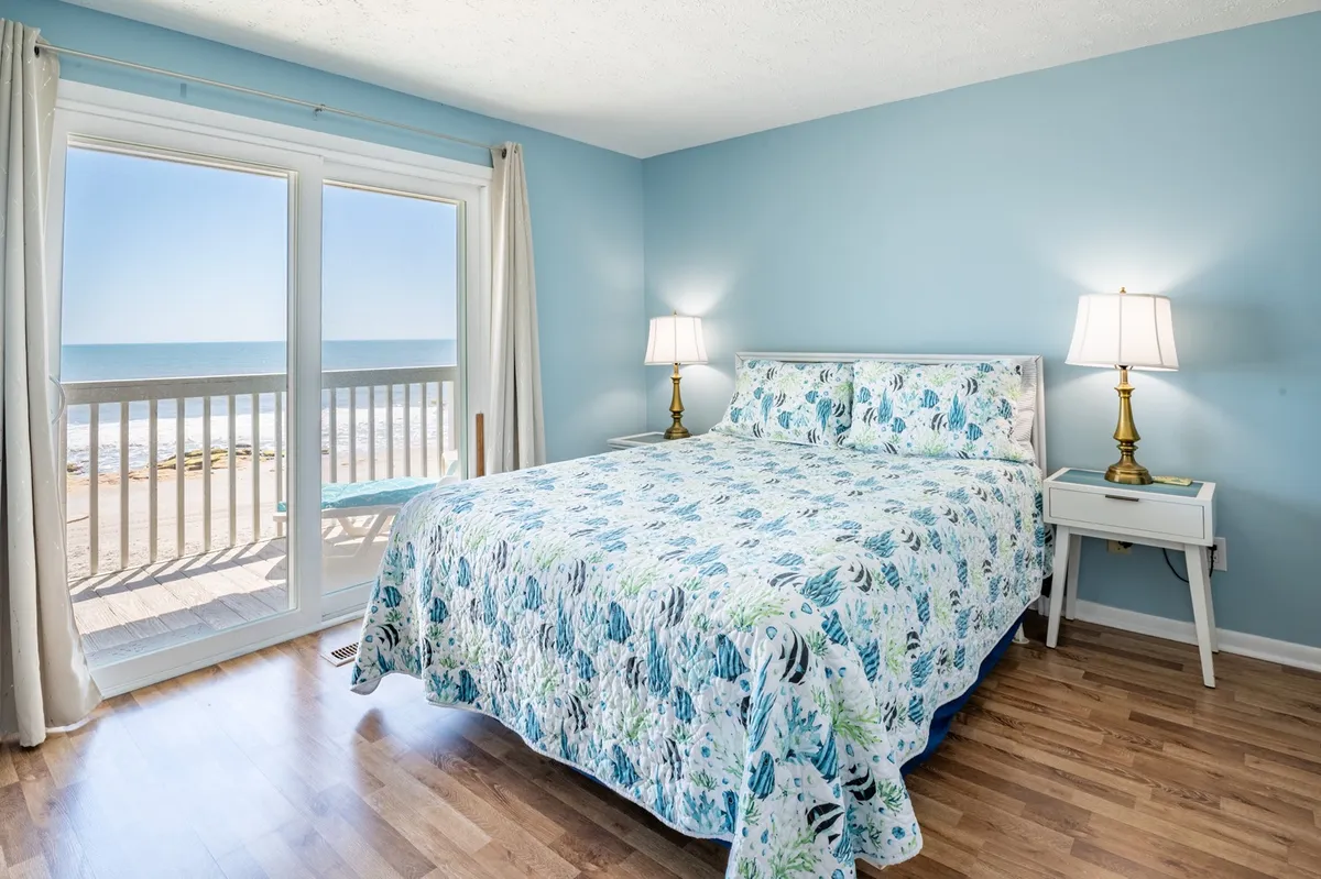 A serene bedroom with floral bedding is anchored by large windows that frame a mesmerizing ocean view. A cozy nightstand with a lamp sits beside the bed, creating an inviting spot for relaxation or reading. Soft natural light fills the room, enhancing its calm, restful atmosphere.