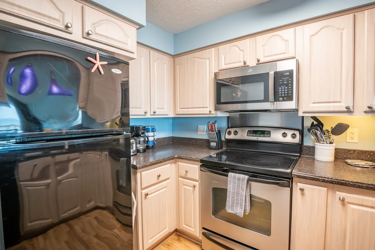 A cozy, fully equipped kitchen with a black refrigerator, stainless steel oven, light wood cabinetry, blue accent walls, and charming starfish decoration. The setup invites meal preparation, family gatherings, and daily routines in a welcoming environment.