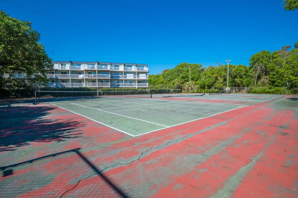 An exterior view of a tennis court surrounded by greenery and an apartment building in the background. The scene captures the energy of outdoor sport and community engagement, with a well-maintained court inviting guests to play or spectate.