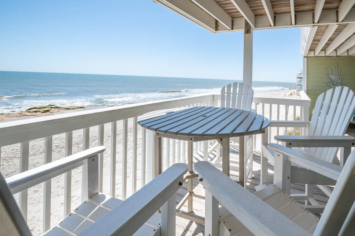 A serene balcony scene overlooking the Atlantic Ocean, featuring two white Adirondack chairs and a wooden table—perfect for enjoying coffee at sunrise or drinks at sunset. The expansive ocean view immediately draws the eye, with the soothing blue water and sky setting a romantic, tranquil tone.
