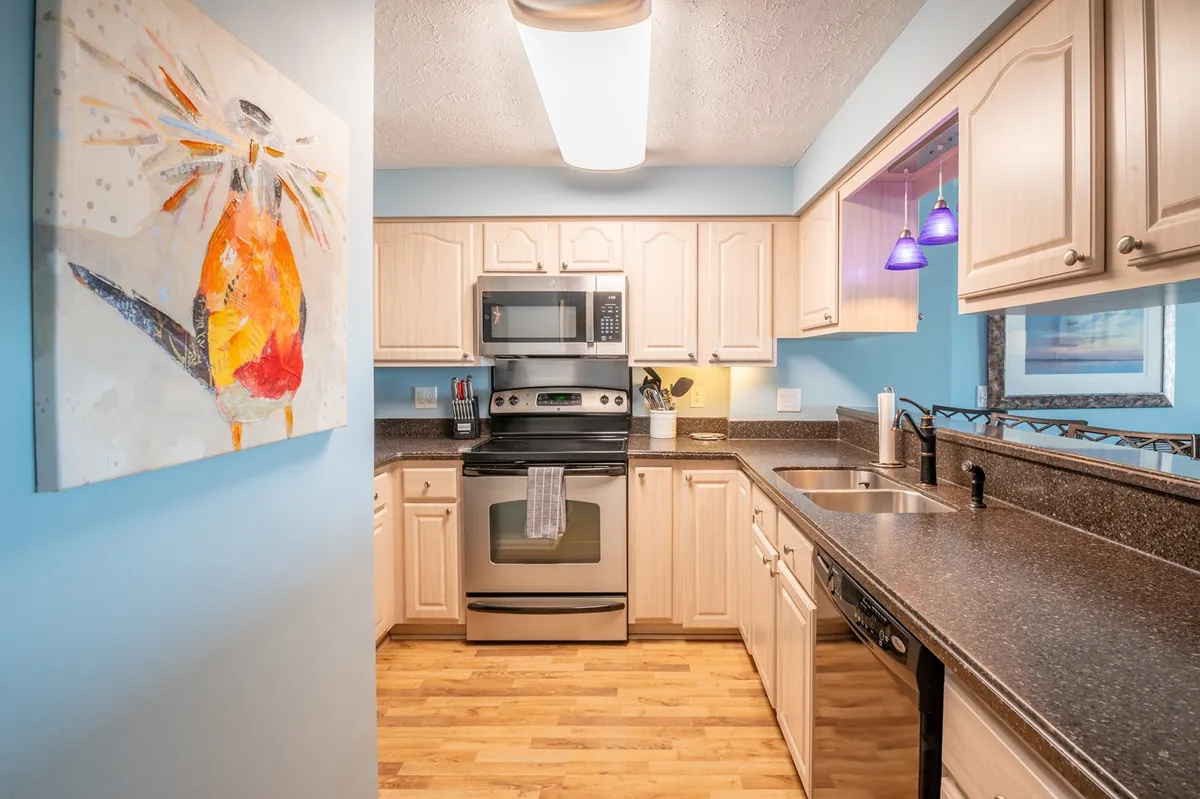 A bright kitchen scene with stainless steel appliances, colorful artwork, ample counter space, and wooden cabinetry, suggesting a practical space for cooking and entertaining. The welcoming vibe highlights the convenience of home-cooked meals and easy living.