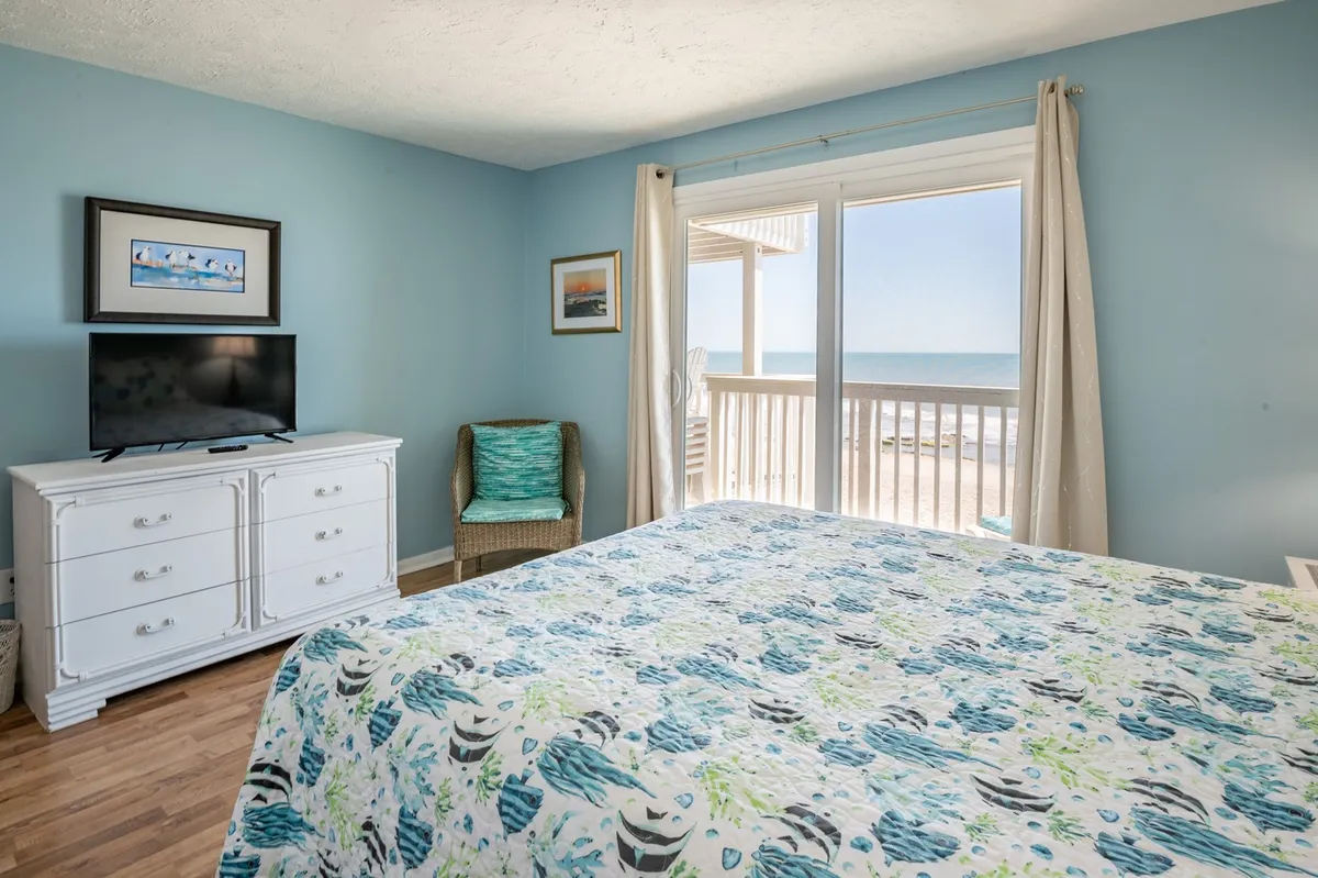 A bedroom featuring a bed with colorful yet soothing bedding, a large window offering ocean glimpses, and a TV unit positioned for convenient viewing. The soft, natural palette and abundance of light create a restful, modern ambiance ideal for unwinding after a day at the beach.