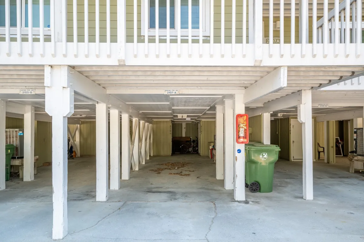 This image displays the parking area beneath the building, featuring visible support beams, a green trash bin, and ample open space for vehicles. The practical layout highlights the convenience of parking close to the unit, emphasizing accessibility and organization for guests.