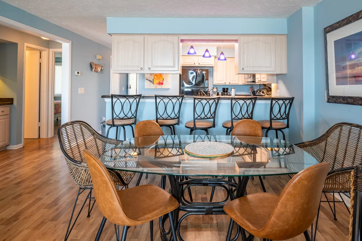 A bright, inviting dining area with a modern glass table and woven chairs, seamlessly connected to an open kitchen. The space is designed for shared meals and casual conversations, with plenty of natural light and a welcoming atmosphere.