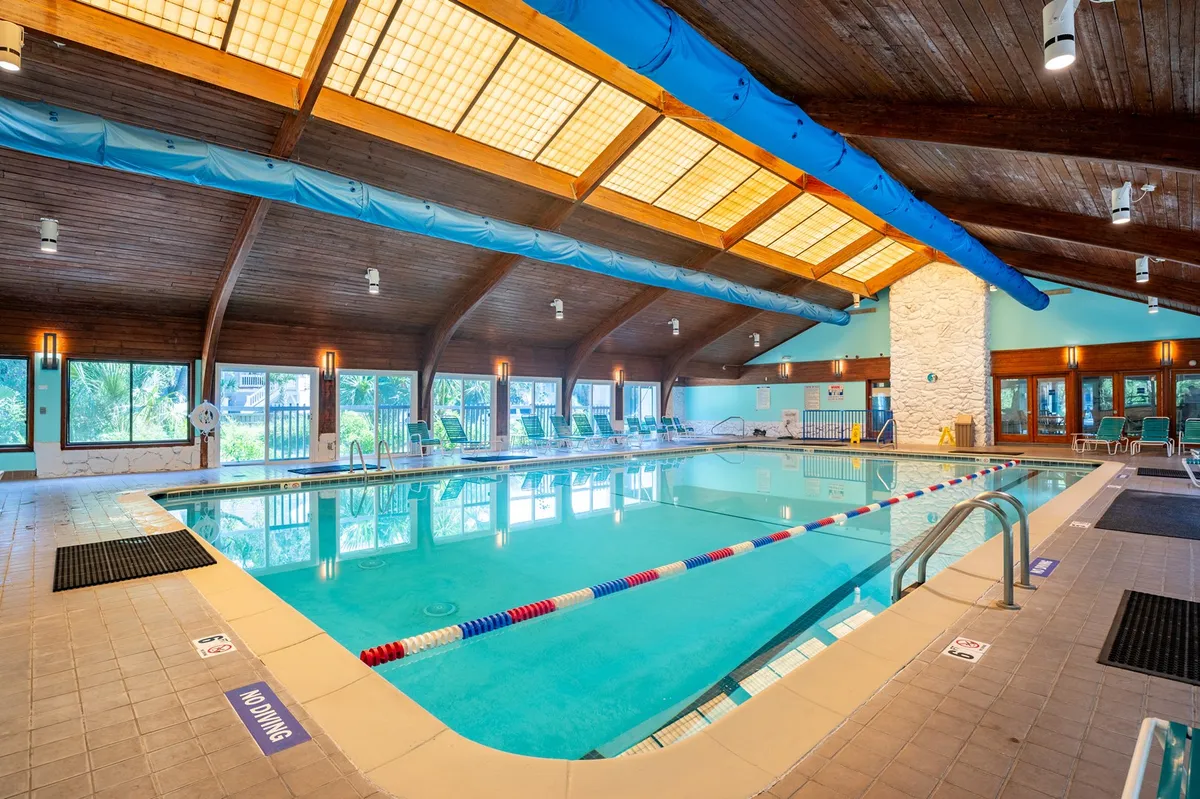 A spacious indoor swimming pool area featuring wooden beams, large windows letting in plenty of natural light, and comfortable poolside loungers. The tranquil setting invites both relaxation and active swimming, and the environment feels modern and rejuvenating—an ideal representation of year-round amenities.