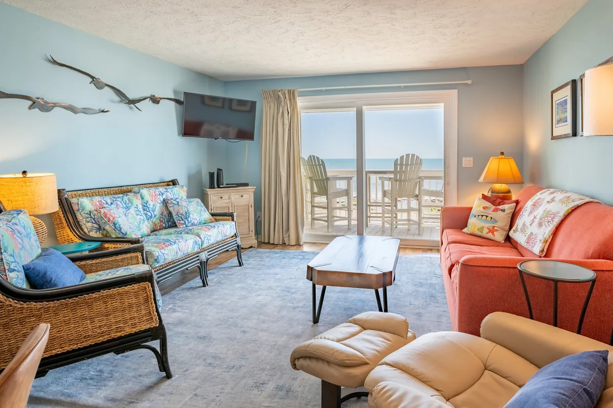 The image captures a bright, cheerful living room with large windows overlooking the ocean. Colorful sofas invite guests to relax, and decorative wall art adds to the coastal ambiance. The wooden coffee table is perfect for gathering around, and the inviting space offers stunning sunrise views over the water.